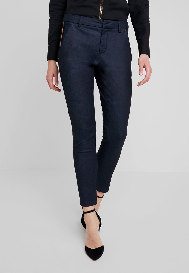 Pantalones ajustados de un oscuro tono navy con una textura suave, que presentan bolsillos laterales con cremallera y una cintura de altura media, combinados con tacones negros de punta afilada.