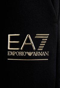 Zwarte stof met een gouden logo "EA7" en "EMPORIO ARMANI" met daaronder een horizontale lijn. Gladde textuur en glanzende accenten.