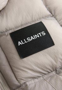 Beige pufferjacka av tyg med matt yta, präglad av en svart rektangulär etikett med "ALLSAINTS" broderat i vitt och synliga sömmar.