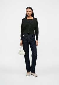 Zwarte knoop-up cardigan, donkerblauwe jeans, beige platte schoenen en een witte geplooide handtas tegen een effen achtergrond.