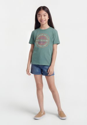 Κορίτσι που στέκεται, φοράει πράσινο t-shirt με κοντά μανίκια και κυκλικό λογότυπο Billabong, τζιν σορτς και καφέ παπούτσια slip-on, χαμογελώντας στην κάμερα.