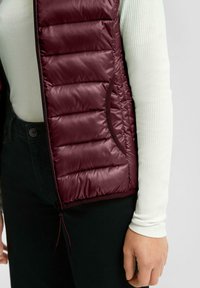 Nahaufnahme einer Person, die eine ärmellose, kastanienbraune gepolsterte Weste über einem weißen, gerippten Langarmoberteil und schwarzen Hosen trägt, wobei die Weste, die Tasche und der Reißverschluss zu sehen sind.