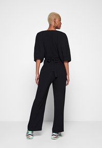 Combinaison noire avec des jambes larges et des manches chauve-souris courtes. Dotée d'une taille à nouer et d'une fermeture éclair dans le dos. Le modèle porte des baskets sportives.