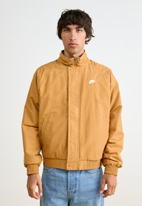 Giacca Nike color sabbia, con colletto alto, chiusura a zip e polsini elasticizzati. Include un logo bianco sul lato sinistro del petto e tasche laterali.