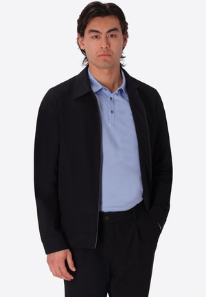 Uomo con capelli scuri che indossa una polo azzurro chiaro, una giacca nera con cerniera e pantaloni neri, in piedi con una mano in tasca.