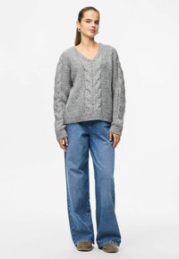 Jeune femme portant un pull en tricot gris à torsades, un jean large bleu et des chaussures brunes, se tenant devant un fond blanc.