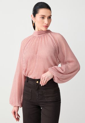 Frau mit dunklen Haaren zu einem tiefen Pferdeschwanz gebunden, trägt eine rosafarbene, strukturierte Bluse mit hohem Kragen und schwarze, hochtaillierte Hose, schaut zur Seite.