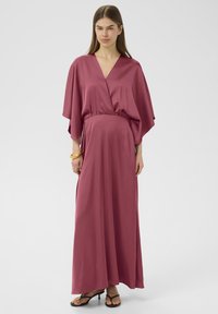 Maxikleid aus weichem, rosafarbenem Stoff mit V-Ausschnitt und weiten Ärmeln, tailliert in der Taille. Kombiniert mit schwarzen High Heels.