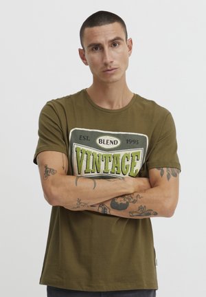 Maslinasto zelena majica od pamučnog materijala s vintage printom loga u bijeloj i zelenoj boji. Okrugli izrez, kratki rukavi i opušteni kroj.