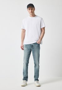 Camiseta de algodón blanca, pantalones vaqueros de mezclilla azul claro y zapatillas blancas. El hombre lleva una gorra negra, de pie frente a un fondo blanco liso.