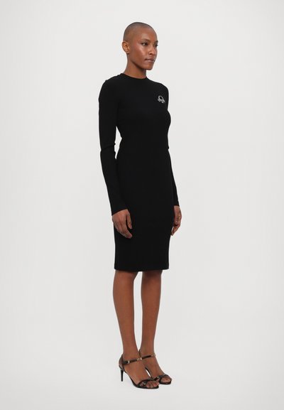 Rochie neagră, cu manșon lung, tip bodycon, având un decolteu rotund și un mic design brodat pe piept, asortată cu tocuri negre cu bretele.