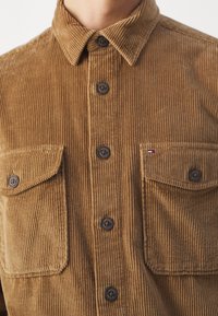 Brun kordfløjlsjakke med vertikale ribbing, spids krave, knapper foran, to brystlommer og kontrasterende mørke knapper. Tekstilen har en blød tekstur.