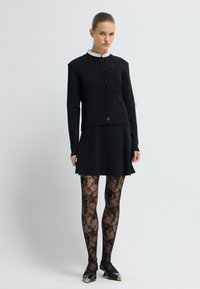 Černá přiléhavá bunda s kulatým límcem a zapínáním na knoflíky, spárovaná s černou sukní. Obsahuje černé punčocháče s krajkovým vzorem a špičaté boty.