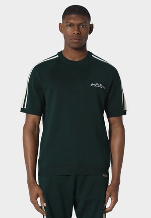 Uomo che indossa un maglione verde scuro a maniche corte con strisce bianche sulle spalle e una piccola scritta bianca sul petto sinistro, in piedi di fronte a uno sfondo semplice.