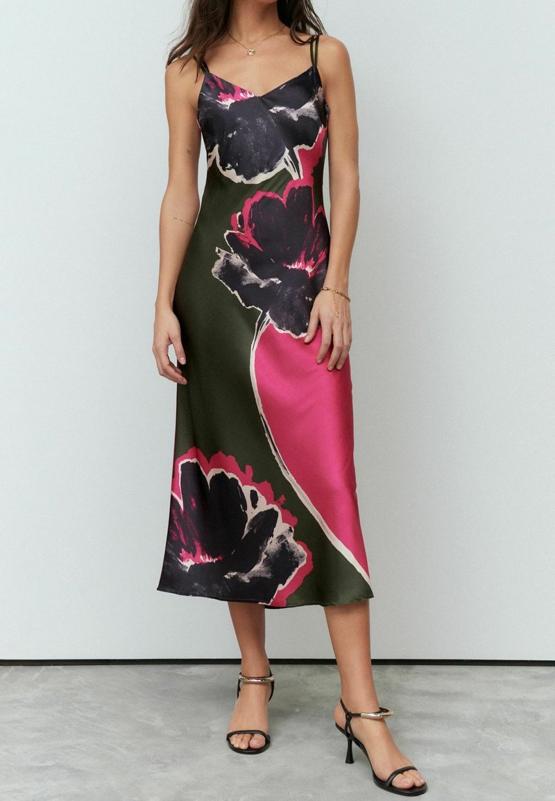 Femme portant une robe midi sans manches avec des motifs floraux vert foncé, rose et noir, associée à des sandales noires à talons hauts.