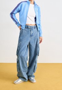 Giacca zip-up azzurro chiaro con dettagli viola, abbinata a pantaloni cargo in denim larghi con tasche e scarpe sportive argentate.