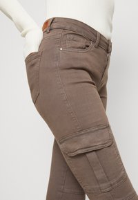 Hnědé cargo kalhoty s úzkým střihem, s bočními kapsami, prošitými detaily a zipovým zapínáním. Vyrobeno z měkké, strukturované tkaniny.