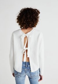 White ribbed knit sweater featuring an open back with tie closures. Paired with fitted light blue jeans, showcasing a casual look.