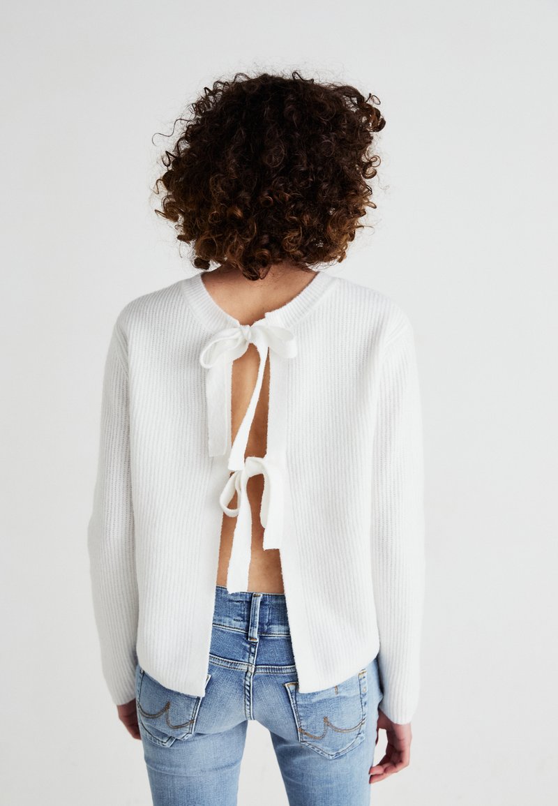 White ribbed knit sweater featuring an open back with tie closures. Paired with fitted light blue jeans, showcasing a casual look.