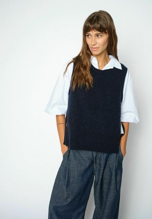 Femme aux longs cheveux bruns portant une chemise blanche sous un pull sans manches noir et un pantalon ample bleu foncé, debout les mains dans les poches.