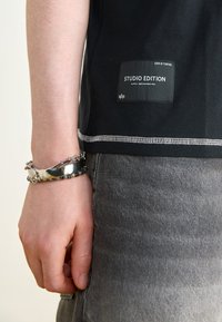 T-shirt noir avec une étiquette "STUDIO EDITION" cousue, associée à un jean gris. Bracelets en argent visibles sur une main reposant sur le côté.