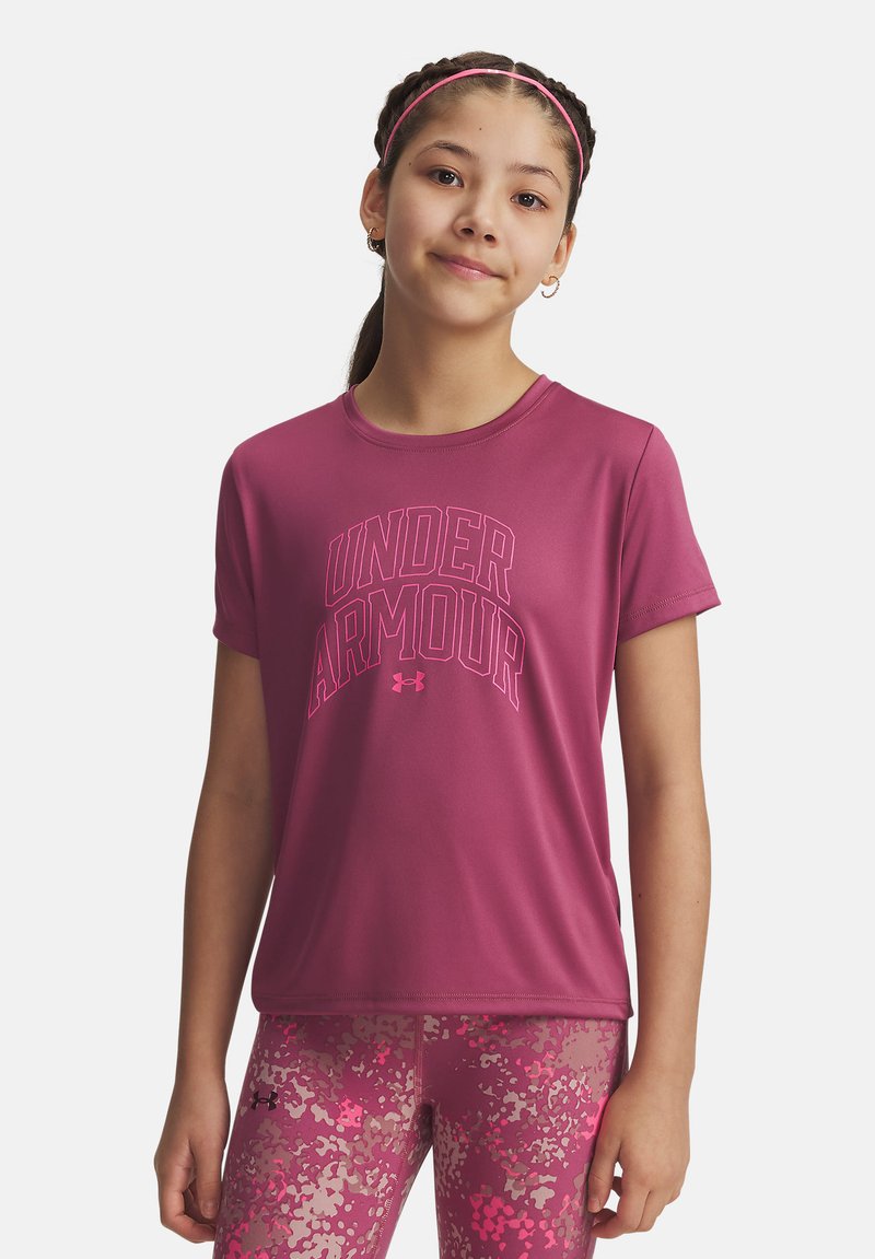 Ragazza che indossa una t-shirt rosa Under Armour e leggings rosa a fantasia, in piedi con un leggero sorriso su sfondo bianco.