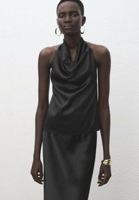 Čierny saténový top na ramienka s drapériou, skombinovaný s dlhou sukňou. Doplnky zahŕňajú zlatý náramok a kruhové náušnice.