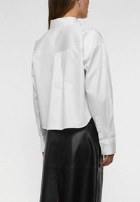 Hvid skjorte med krave, cropped ryg, lange ærmer og manchet med knapper, kombineret med en sort lædernederdel med sideslids.