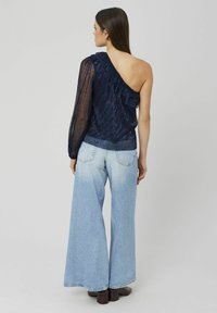 Top asymétrique marine avec détails froncés, manches longues transparentes, associé à un jean à taille haute bleu clair à jambes larges et des bottes marron foncé.