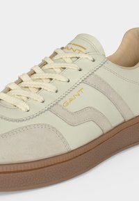 Baskets crème et beige avec semelle marron texturée, lacets beiges et logo "GANT" en or sur le côté et la languette.