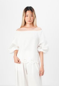 Blusa bianca scollata sulle spalle con maniche a palloncino, caratterizzata da dettagli floreali ricamati nell'orlo. Realizzata in tessuto leggero.