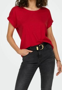 Kvinde iført en rød kortærmet bluse stoppet ned i højtaljede sorte jeans med et sort bælte med en guldbuckle.