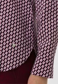 Chemise à manches longues avec un motif géométrique noir, blanc et rouge. Présente un ourlet arrondi et des poignets boutonnés, fabriquée dans un tissu léger.