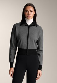 Czarna i szara żebrowana kurtka na zamek z wysokim kołnierzem, dopasowanym pasem i teksturowaną tkaniną, zestawiona z czarnymi legginsami.