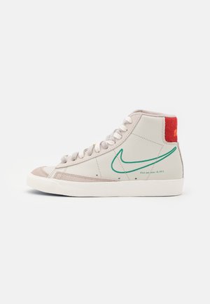 Beżowy wysoki sneaker z białymi sznurowadłami, zielonym konturem logo, czerwoną pętelką na pięcie i tekstem "Pierwsze użycie 18 czerwca 1971" po boku.