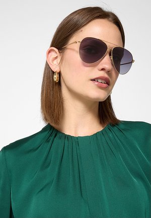 Donna con capelli castani lisci indossa grandi occhiali da sole con montatura dorata, orecchini a cerchio dorati e una camicetta verde scuro plissettata.