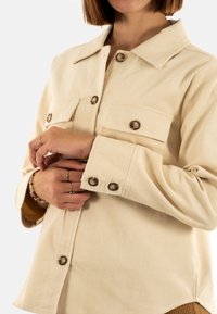 Chaqueta de pana beige con cuello, dos bolsillos en el pecho y botones marrones. Presenta un tejido texturizado y mangas largas con puños abotonados.