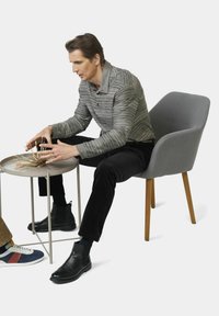 Graues gestreiftes Langarmshirt, schwarze Cordhosen und schwarze Chelsea-Stiefel. Sitzt an einem hellgrauen Beistelltisch mit bunten Mikadostäbchen.