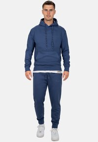 Felpa blu navy con tasca frontale e coulisse, abbinata a pantaloni sportivi coordinati con polsini elasticizzati e un patch con logo.