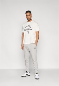 T-shirt branca de manga curta com letras escuras, calças de treino cinzentas e ténis brancos. Apresenta o logótipo e o texto da Seleção Nacional de Futebol Masculino dos EUA.