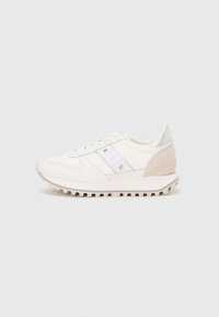 Des baskets athlétiques blanches avec une tige en tissu texturé, des accents argentés et un talon en daim beige. La semelle présente un design rainuré pour une meilleure adhérence.