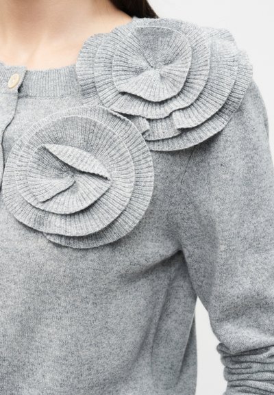 Γκρι πλεκτό πουλόβερ με δύο μεγάλες, υφασμάτινες διακοσμήσεις με σπειροειδές μοτίβο που μοιάζουν με λουλούδια στον ώμο και την περιοχή του στήθους κοντά σε λαιμόκοψη με κουμπιά.