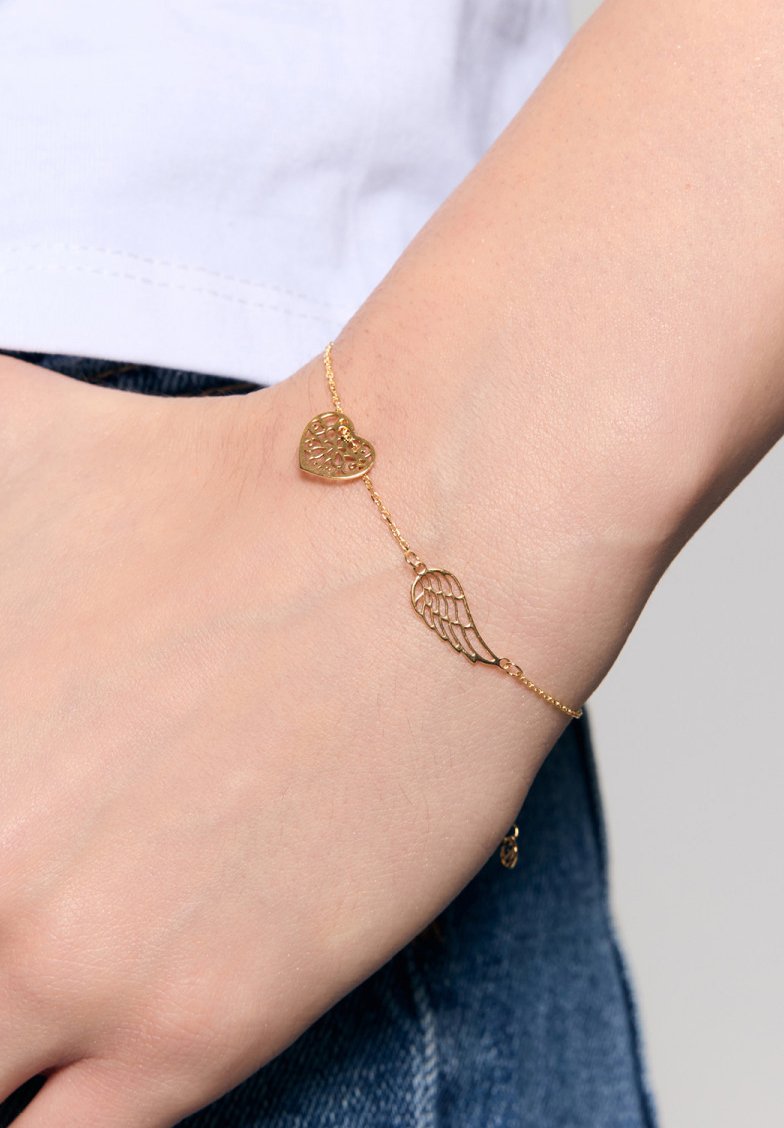 Złota bransoletka z sercem i skrzydłem, ozdobiona misternymi wzorami wyciętymi w materiale. Cienki łańcuszek o delikatnej teksturze noszony na nadgarstku.