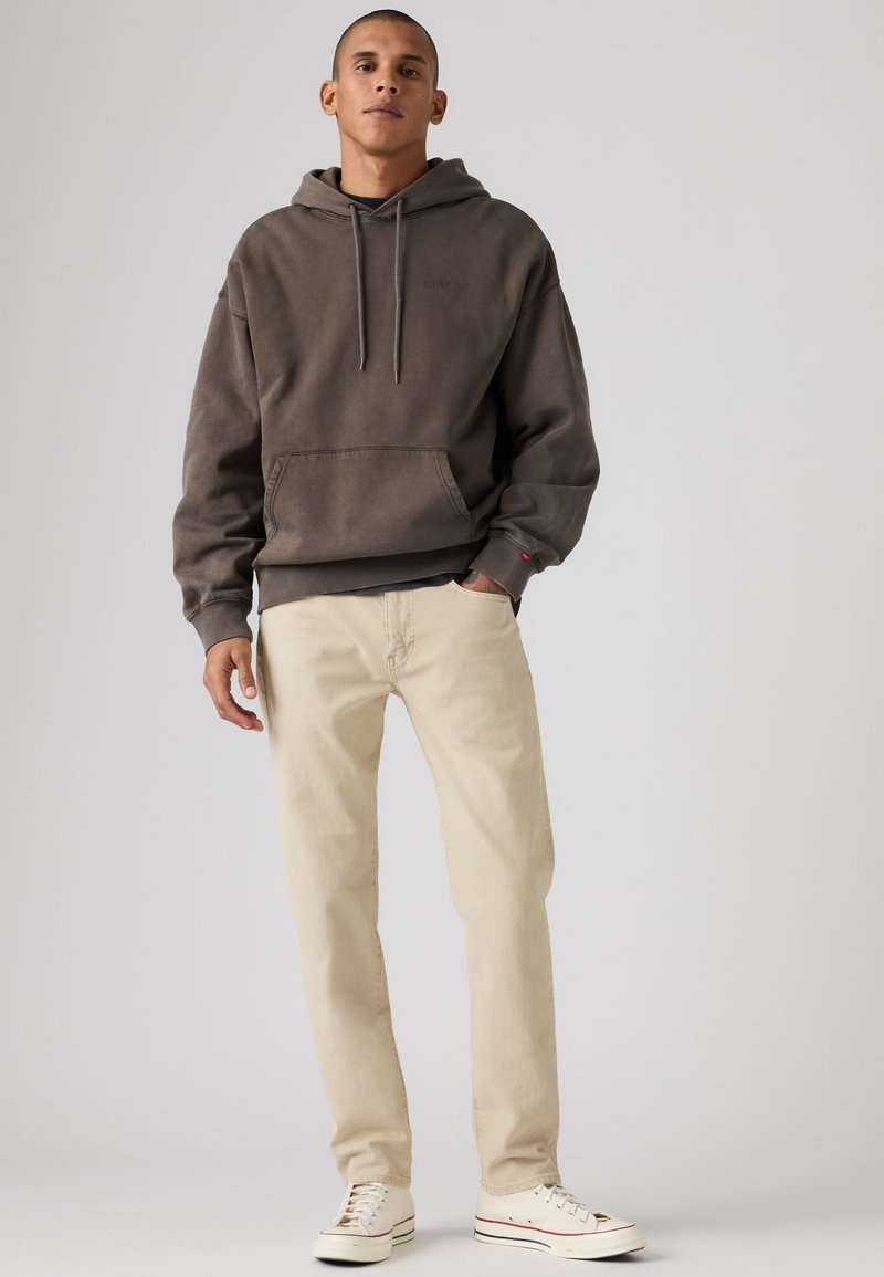 Uomo che indossa una felpa con cappuccio marrone, pantaloni beige e sneaker bianche, in piedi con una mano in tasca contro uno sfondo chiaro e uniforme.