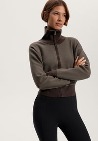 Brown zip-up sweatshirt with a high collar, ribbed texture, and contrasting lower band. Worn over black fitted leggings.