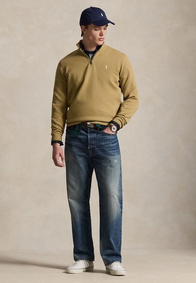 Jaunas vyras dėvi šviesiai rudos spalvos pusiau užtraukiamą megztinį, išeikvotus mėlynus džinsus, baltus sportinius batelius ir tamsiai mėlynę kepurę, stovintis su ranka kišenėje.