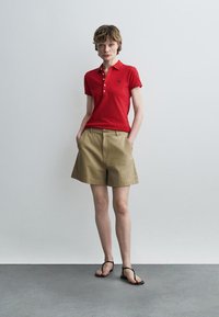 Ung person stående iført en rød kortærmet polo, beige shorts med brede ben og sorte sandaler med remme mod en ensfarvet baggrund.