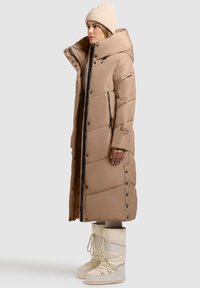 Hosszú, bézs színű puffermellény magas gallérral, karcsúsított fazonnal és nagy gombokkal. Krém színű hócsizmával és bézs kötött sapkával párosítva.