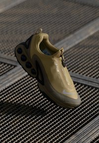 Beige and black athletic shoe with zipped closure and visible air cushion sole, displayed above textured metal grate flooring.