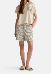 Žena v béžovém tričku s krátkým rukávem henley, vzorovaných béžových a hnědých šortkách, šátku s třásněmi a béžových nazouvacích sandálech se zkříženými popruhy.